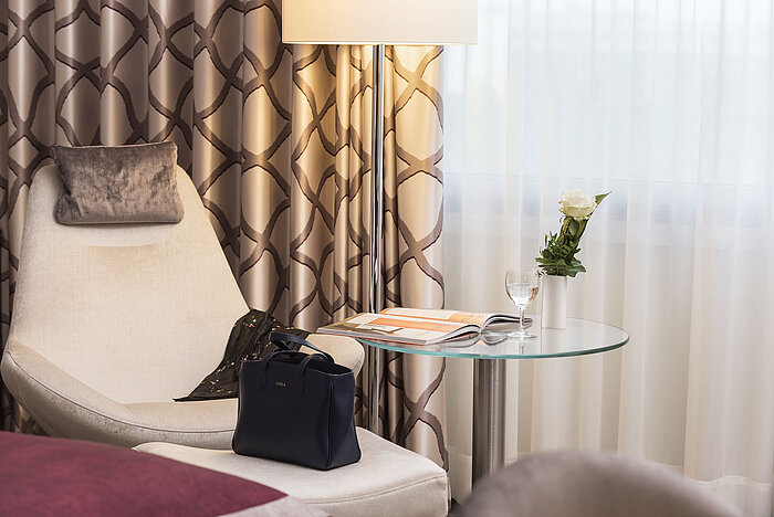 Detail of a Classic room at Maritim Hotel Bonn with a cozy chair, curtain, table, glass, rose, and a handbag.