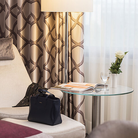 Detail of a Classic room at Maritim Hotel Bonn with a cozy chair, curtain, table, glass, rose, and a handbag.