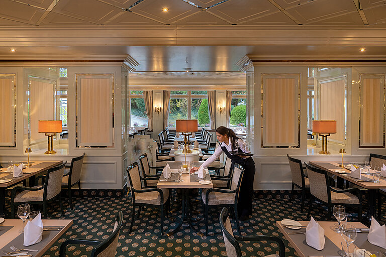 Maritim Restaurant Elegant restaurant with set tables, large windows and a staff member preparing the dining area.