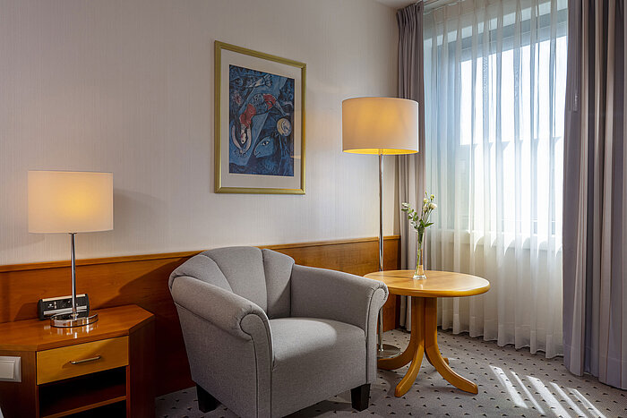 Classic room Bright hotel room in the Maritim Airport Hotel Hannover with armchair, lamp, window, and artwork.