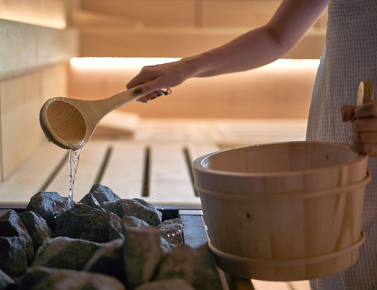 Person pouring water from wooden ladle onto hot sauna stones for infusion