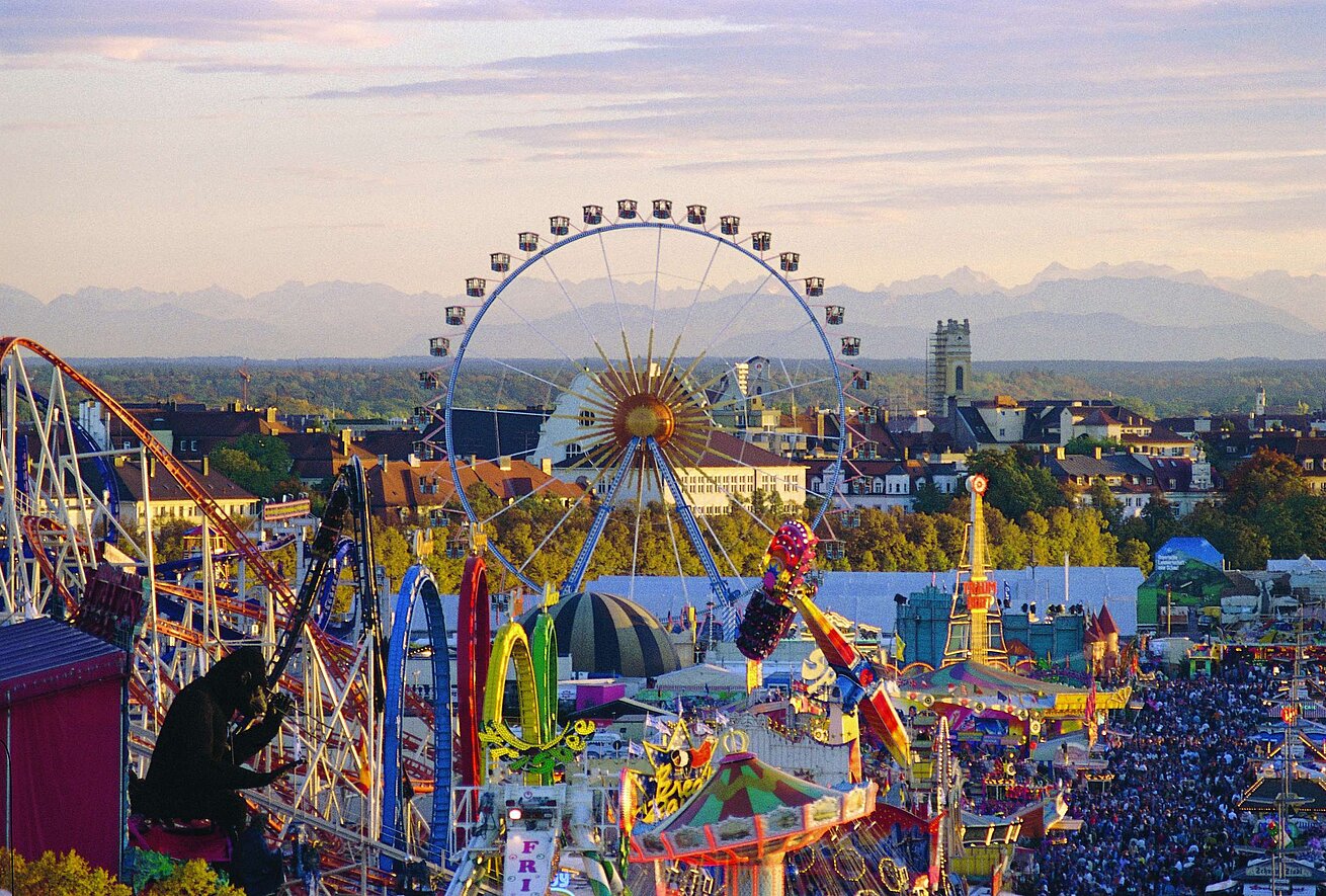 View over the Oktoberfest in Munich: Ferris wheel and rides in front of the city skyline and Alpine scenery.
