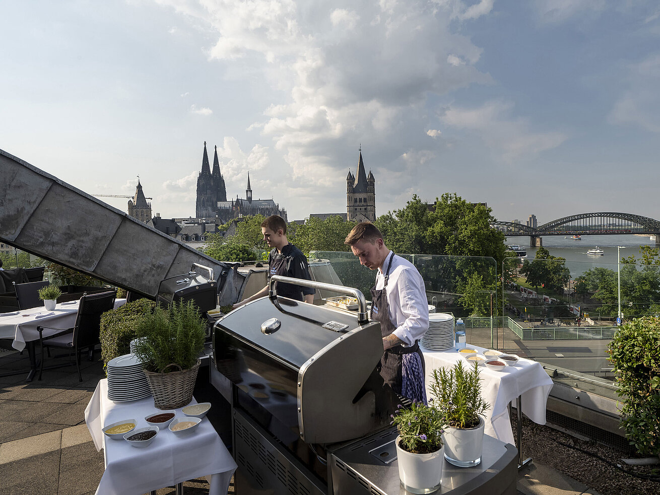 Rooftop terrace of Maritim Hotel Cologne with a view of the cathedral and Hohenzollern Bridge. Chefs preparing a BBQ.