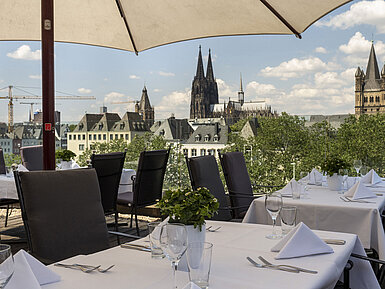Rooftop terrace of Maritim Hotel Cologne overlooking the Rhine and Hohenzollern Bridge, with set tables under umbrellas.